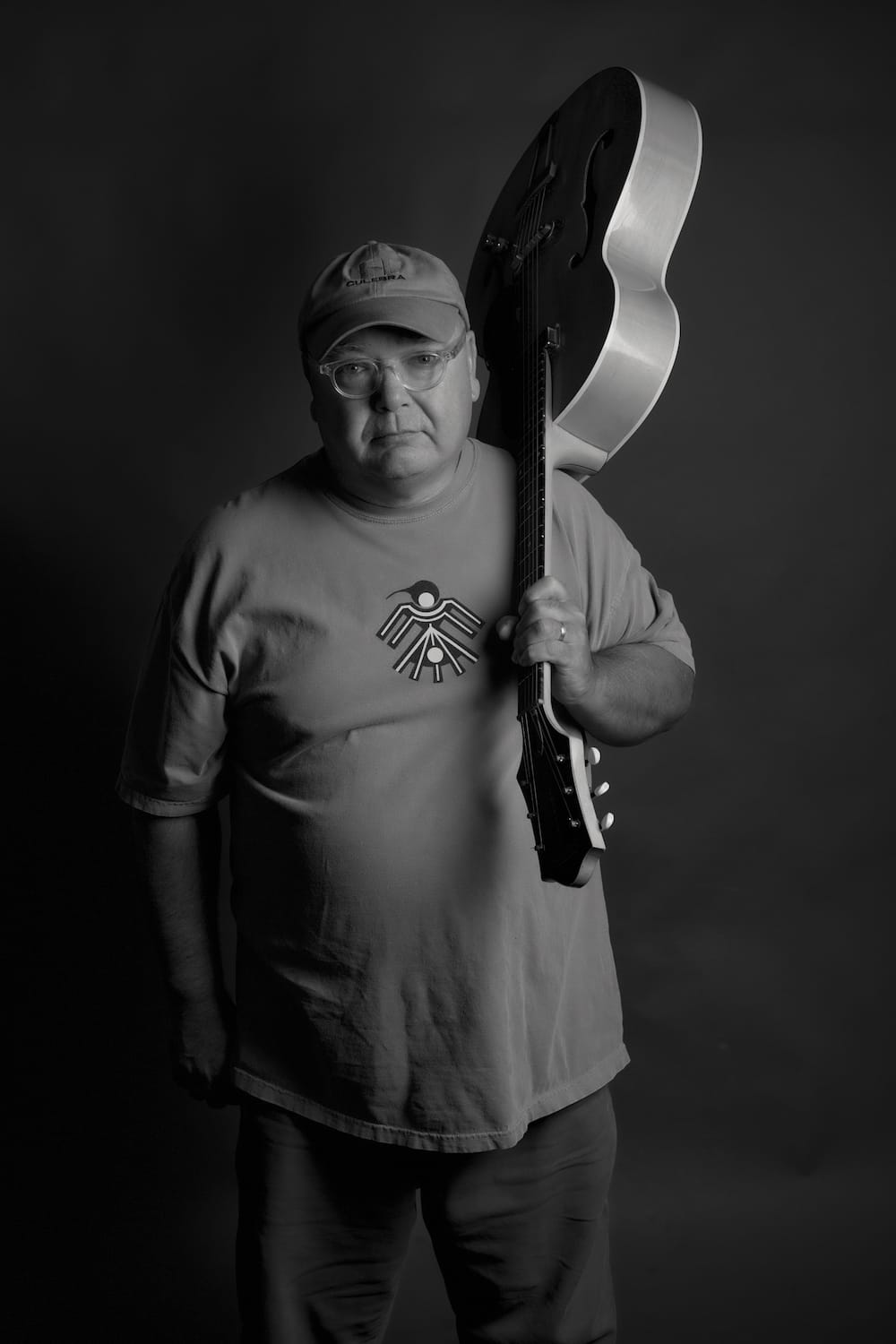 Black-and-white portrait of a man wearing a cap and glasses, holding an acoustic guitar resting on his shoulder in a studio setting.