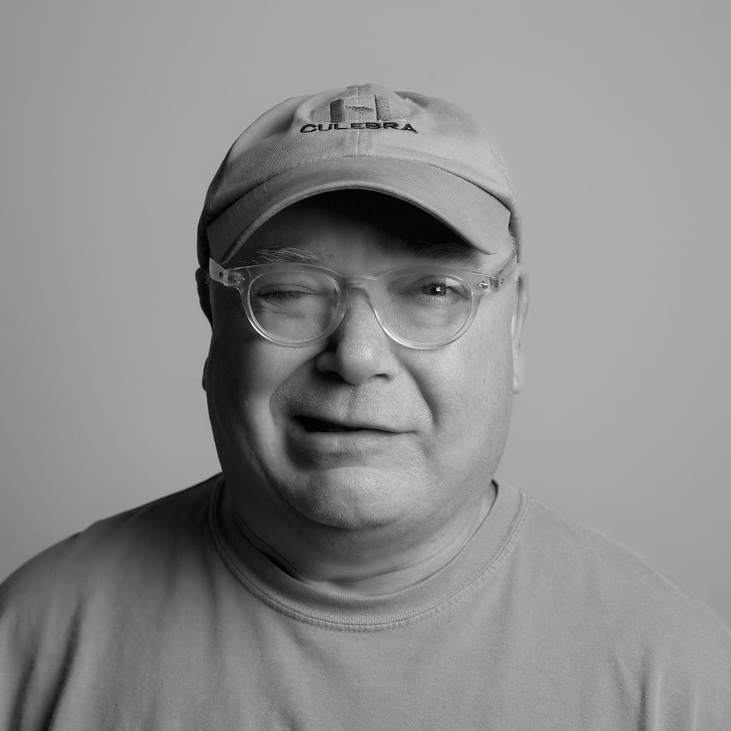 Close-up black-and-white portrait of a man wearing a baseball cap with a CULEBRA logo and clear-framed glasses, smiling softly.