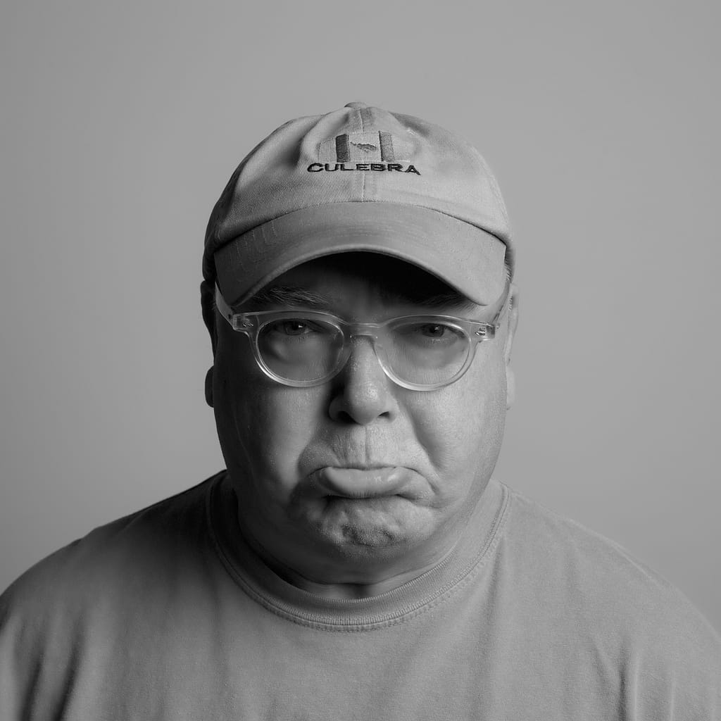 Black-and-white portrait of a man wearing a light baseball cap with 'CULEBRA' logo and clear glasses.