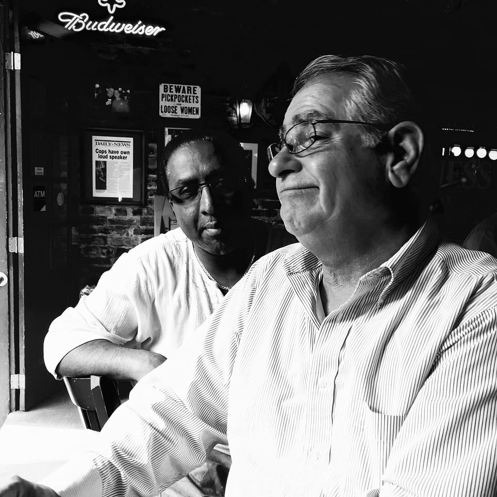 Two men in a black-and-white scene, one wearing glasses in the foreground, with a brick wall and posters behind including a 'BEWARE PICKPOCKETS' sign and a Budweiser neon sign.