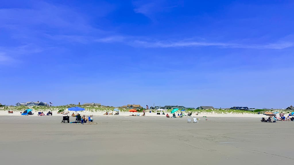 Img_0497 | David Todd Mccarty Wide beach under a bright blue sky with groups of people, colorful umbrellas, and houses along the dunes in the distance.