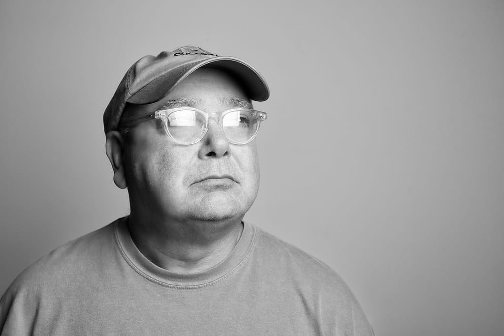 Black-and-white portrait of a middle-aged man in a cap and clear glasses, looking to the right.