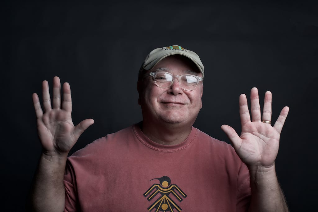 Smiling man wearing a beige cap and clear glasses, waving with both hands against a dark backdrop.