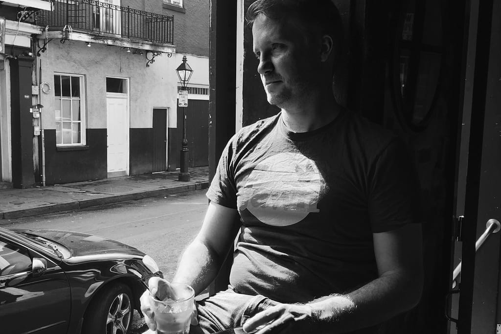 Man in a dark T-shirt sits by a street side, holding a glass with ice, looking to the left; parked car and urban buildings behind him (black-and-white).
