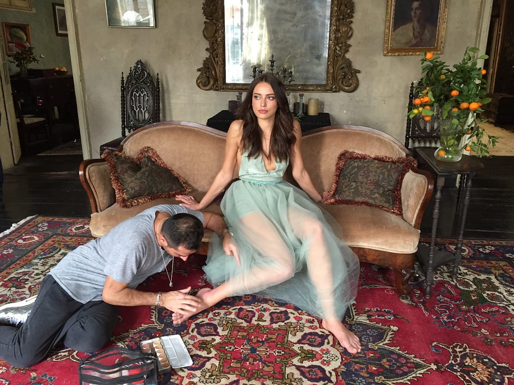 Woman in a mint dress sits on a vintage sofa as a man applies makeup to her foot on a patterned rug in a historic living room with ornate decor.