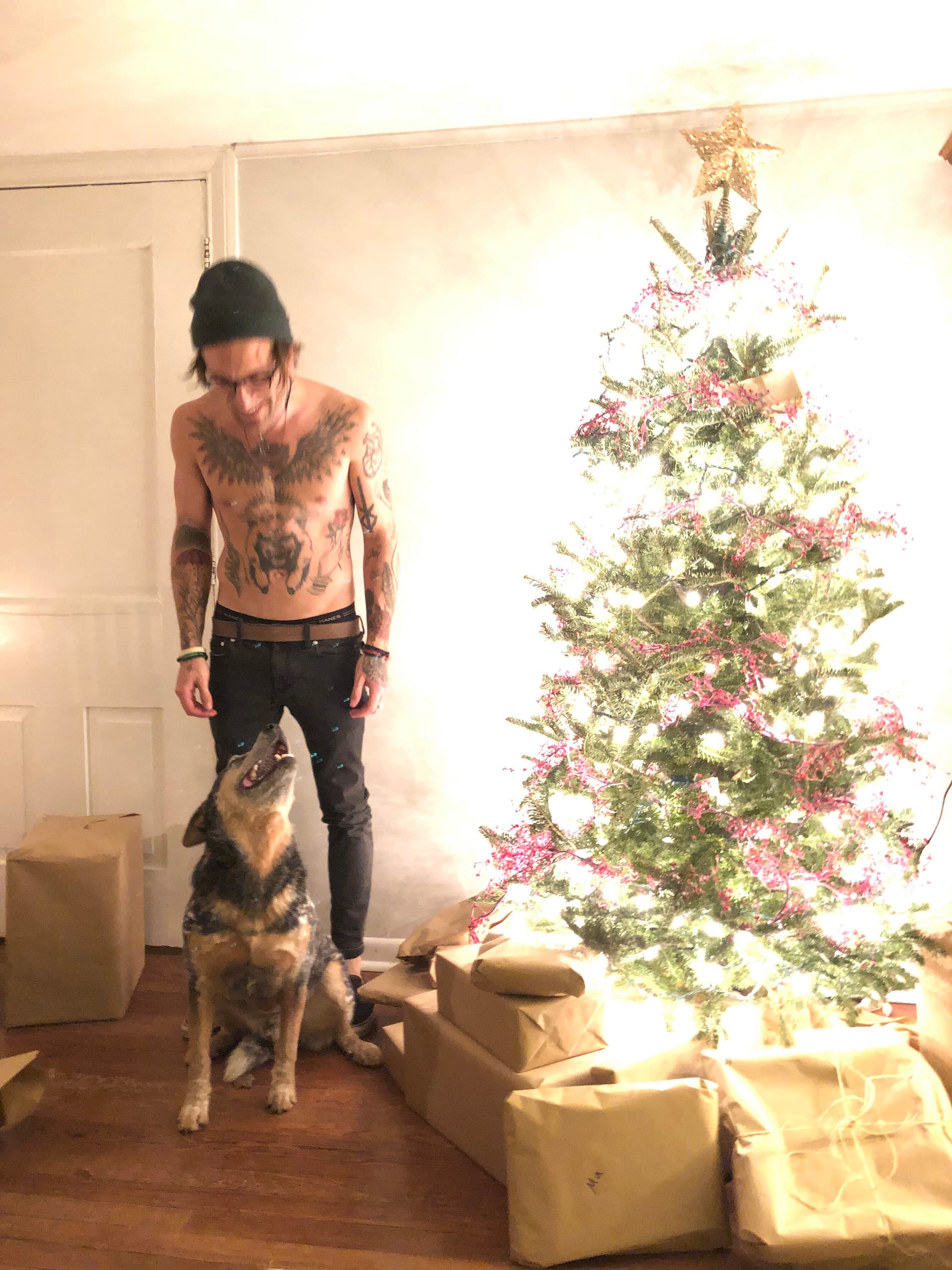 Img_0622 | David Todd Mccarty Tattooed shirtless man wearing a beanie stands next to a dog in a living room with a decorated Christmas tree and wrapped presents nearby.