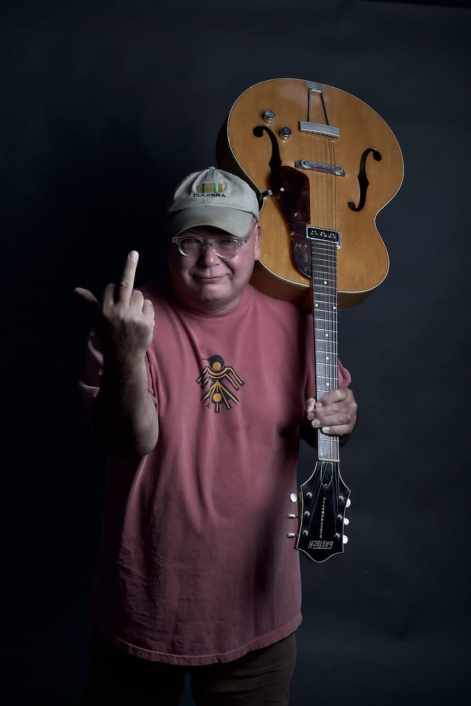 Man wearing a pink t-shirt and cap holds an acoustic guitar over his shoulder while flashing a middle finger at the camera.