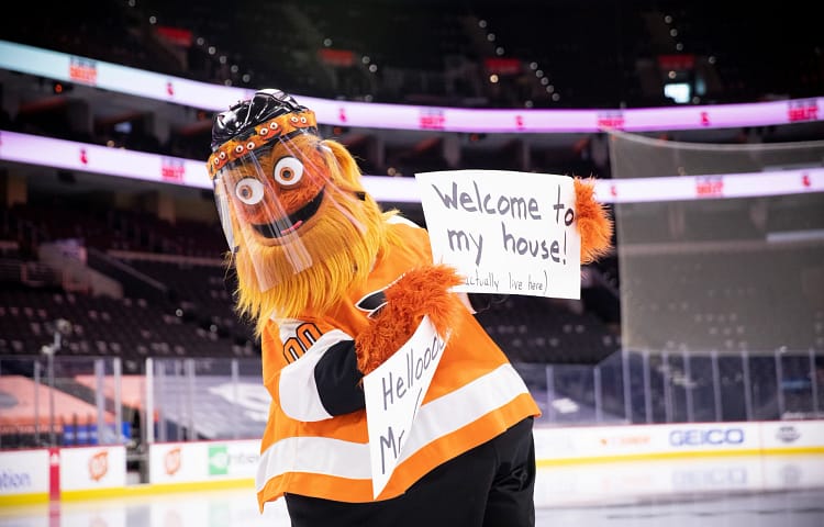 Costumed orange mascot wearing a face shield and orange-and-black jersey holds a sign reading “Welcome to my house!” at an ice hockey arena.