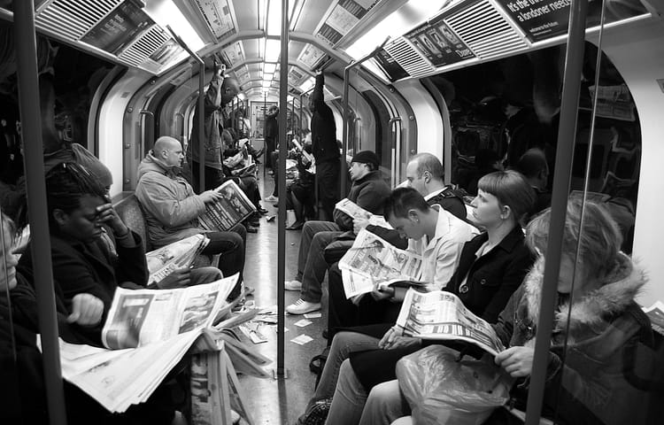 Commuters sitting and standing in a subway car, all reading newspapers.