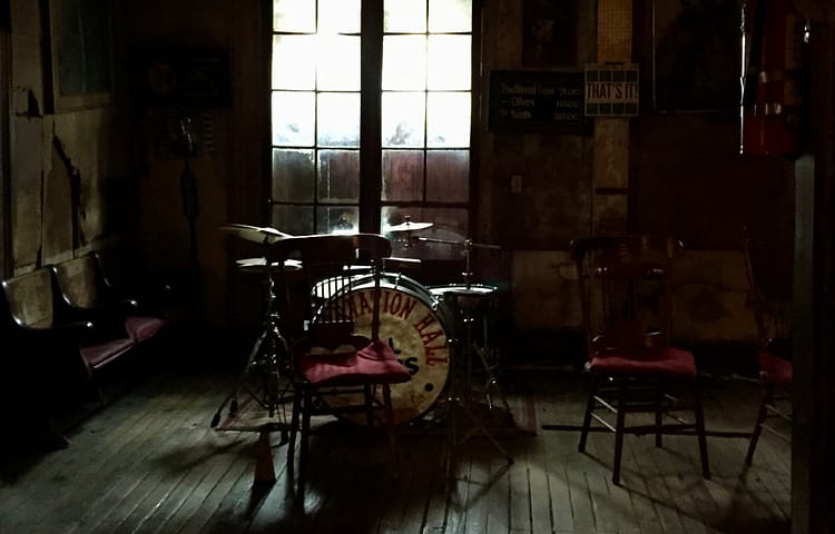 Dimly lit room with a drum kit on a small stage, backlit by large windows behind it, and wooden chairs around the space.