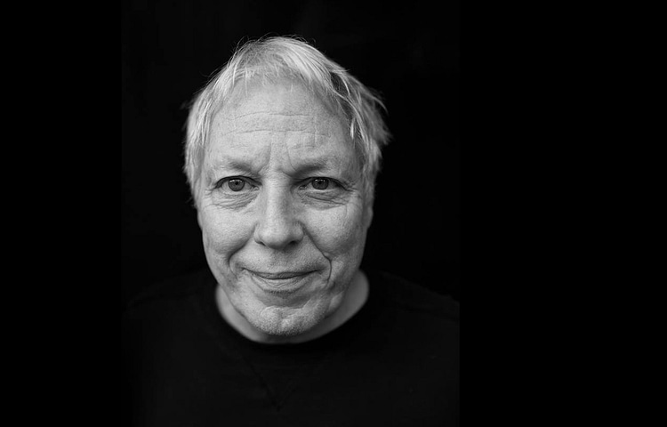 Close-up black-and-white portrait of an older man with light hair and a gentle smile, against a dark background.