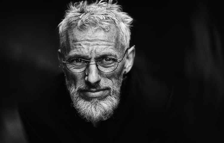 Close-up black-and-white portrait of an older man with gray hair, beard, and round glasses looking at the camera.