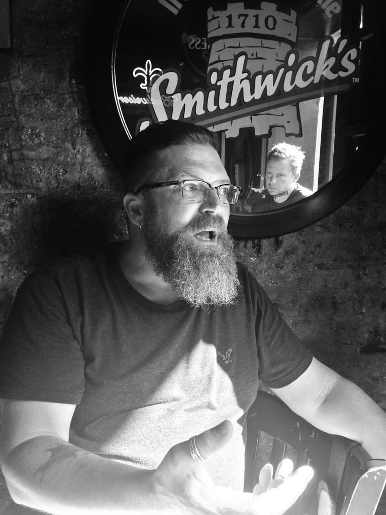 Bearded man with glasses playing a guitar in front of a circular Smithwick's sign (black and white photo).