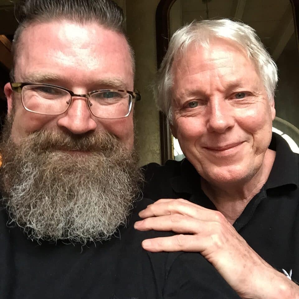 Two men take a close-up selfie, one with a full gray beard and glasses, the other with white hair, both smiling at the camera.