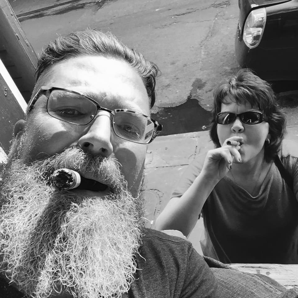 Man with a thick gray beard and glasses smoking a cigar, taking a selfie outdoors.