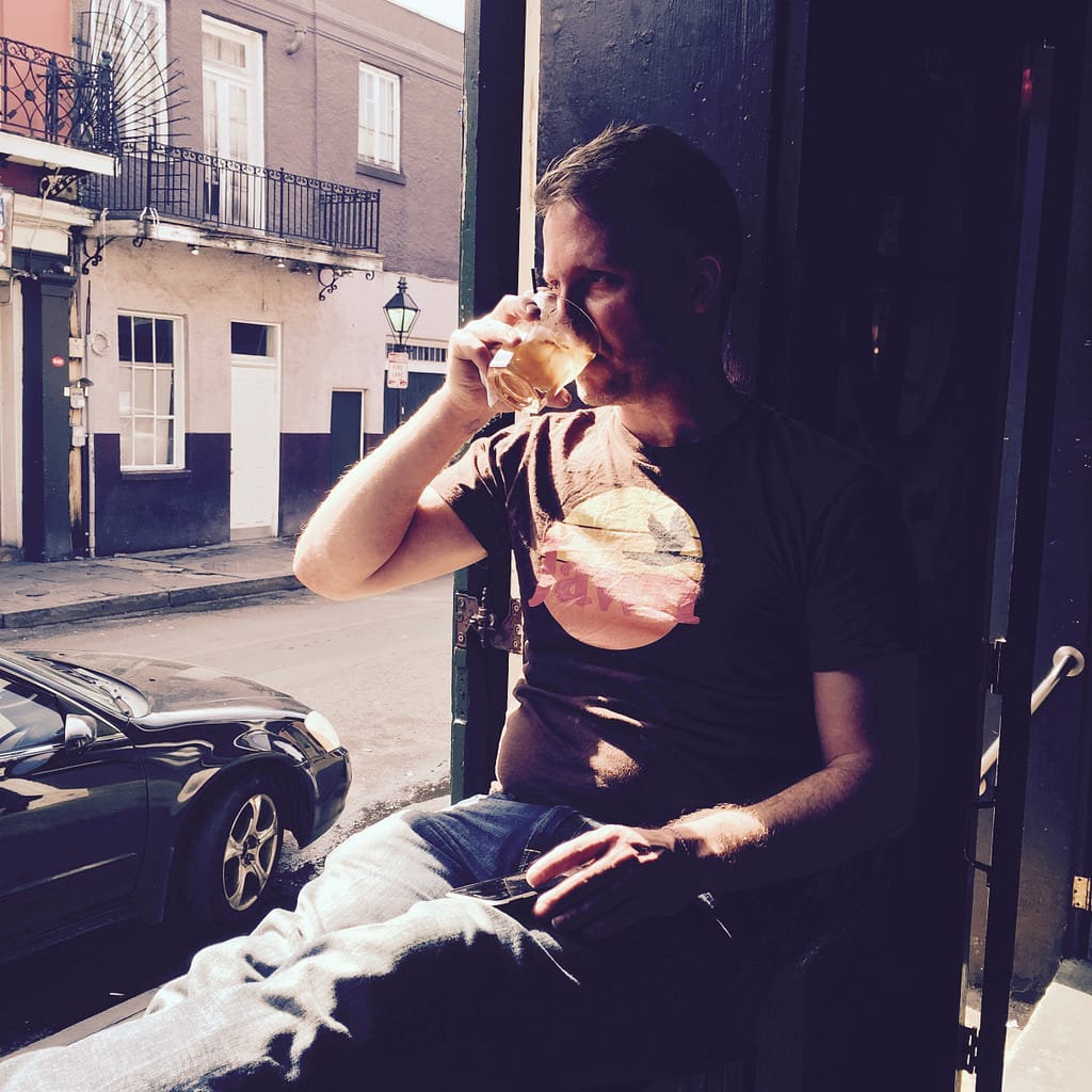 Man in a brown T-shirt sitting by an open door, drinking from a glass outside on a sunny urban street.