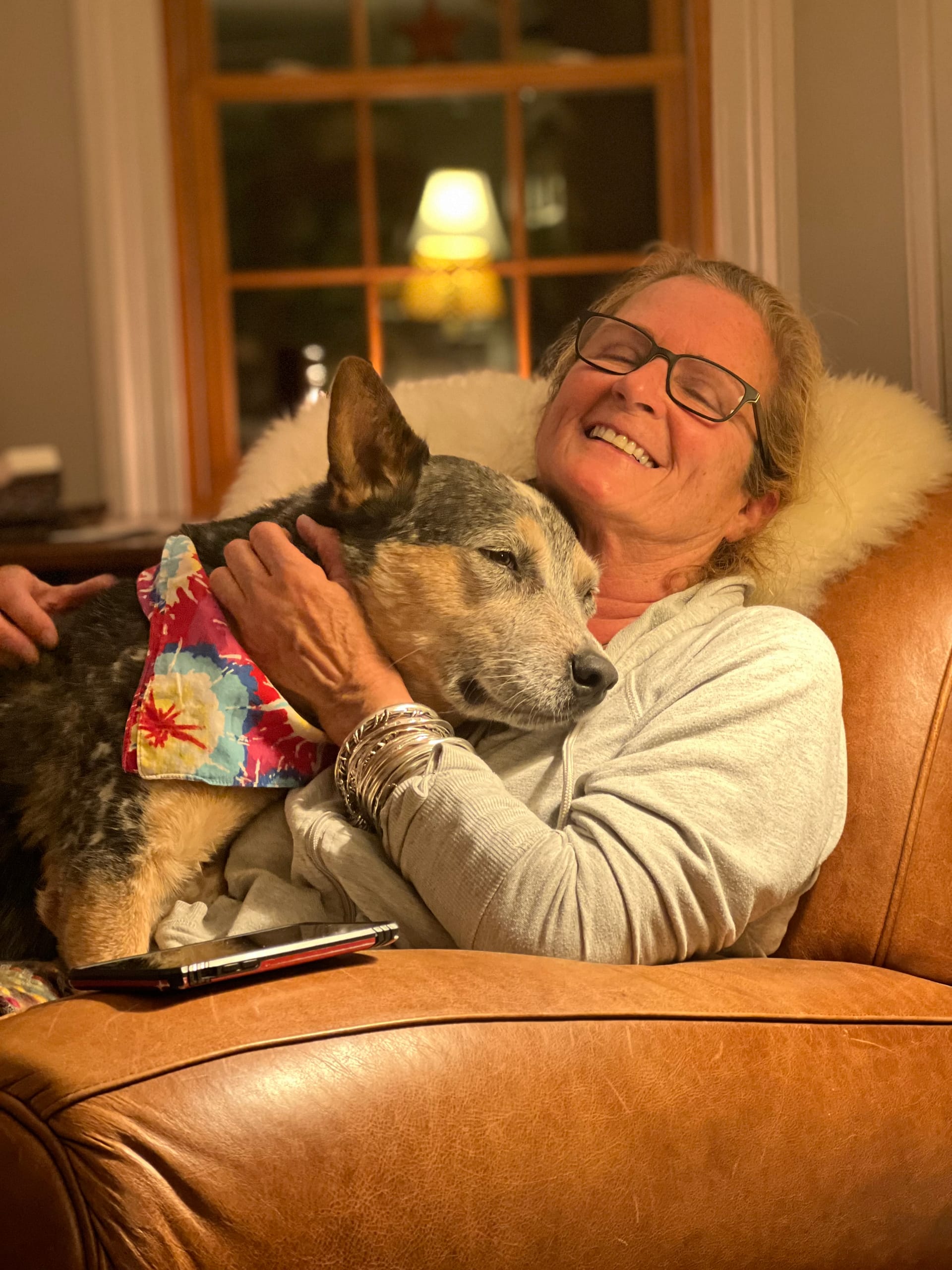 Img_1106 | David Todd Mccarty Smiling woman wearing glasses hugs a dog on a brown leather couch by a window. A colorful bandana rests on the dog's neck.
