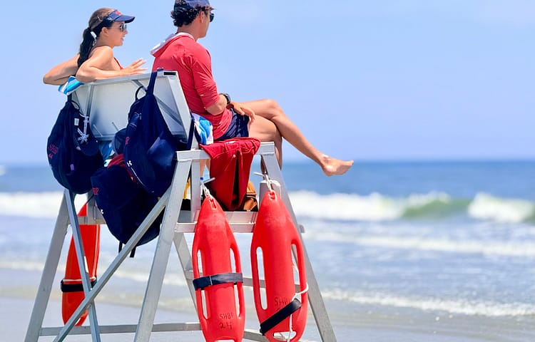 Two lifeguards sit on a wooden lifeguard tower by the ocean, with red rescue tubes hanging from the railing.