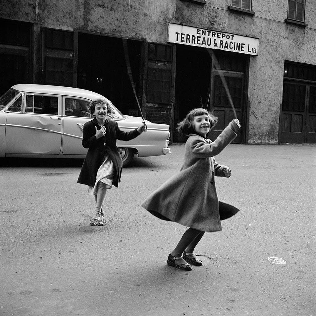 Two young girls skip rope in a street, smiling, beside a vintage car and an old storefront in black and white.