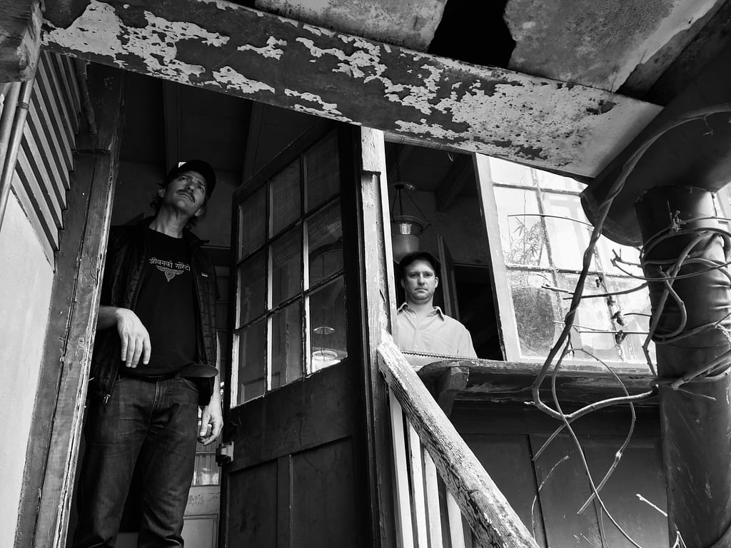 Two men stand in a ruined storefront doorway with peeling paint and chipped wood in a dilapidated building, black-and-white photo.
