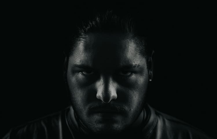 Close-up of a man's face in low lighting, with shadows obscuring most features and a focused, intense gaze from the eyes.