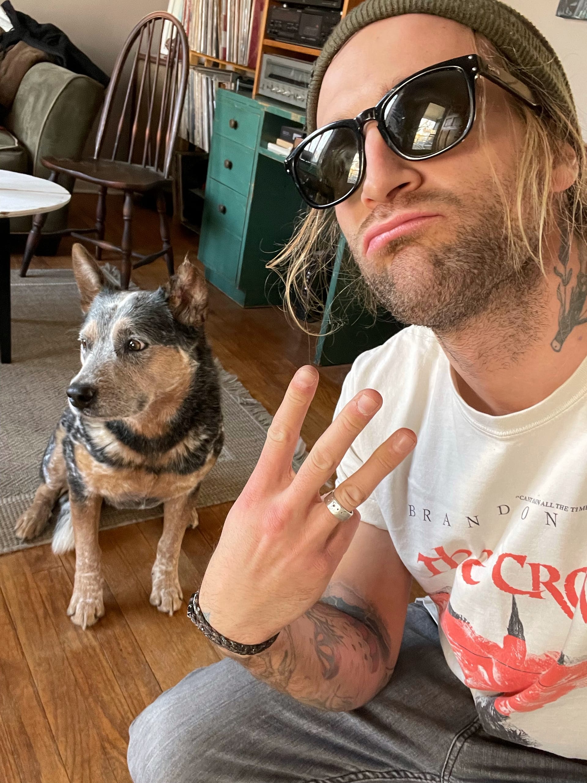 Img_4678 | David Todd Mccarty Man with long hair under a beanie and sunglasses, making a peace sign selfie beside a seated black-and-tan dog in a cozy living room with wooden floor and vintage furniture.
