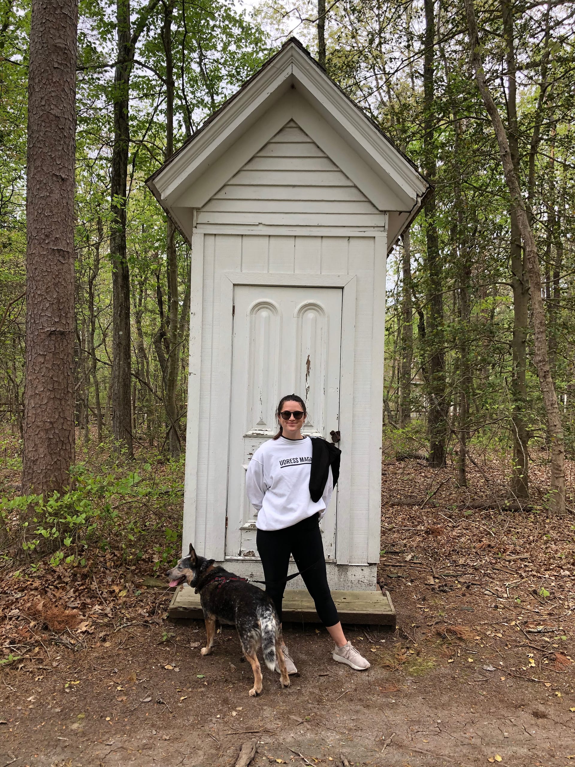 Img_5237 | David Todd Mccarty Woman in a white sweatshirt and sunglasses stands in front of a small white wooden shed in a wooded area, with a dog on a leash nearby.