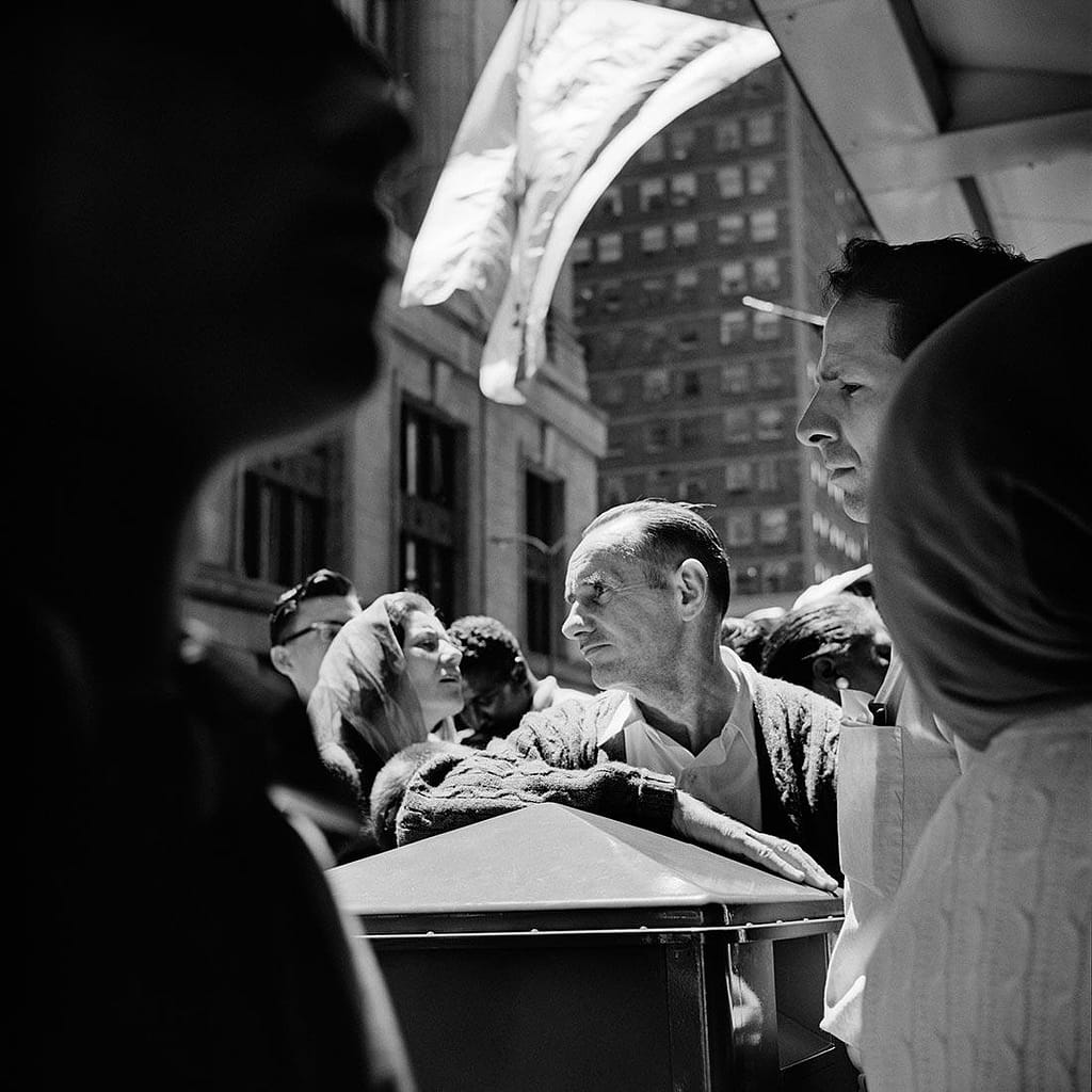 A somber man rests his arm on a dark casket in a crowded urban crowd, black-and-white street scene inviting condolence or mourning.