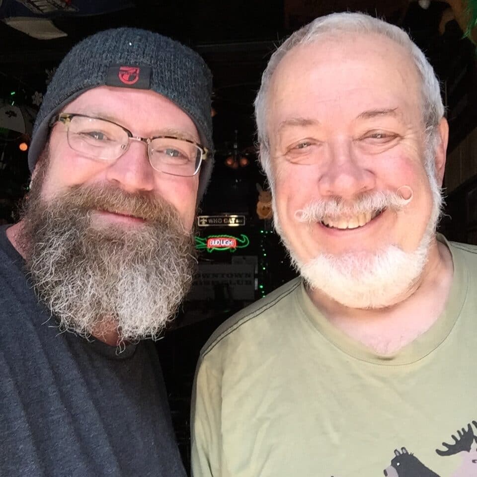 Two men with beards smiling for a selfie, indoors in a casual setting.