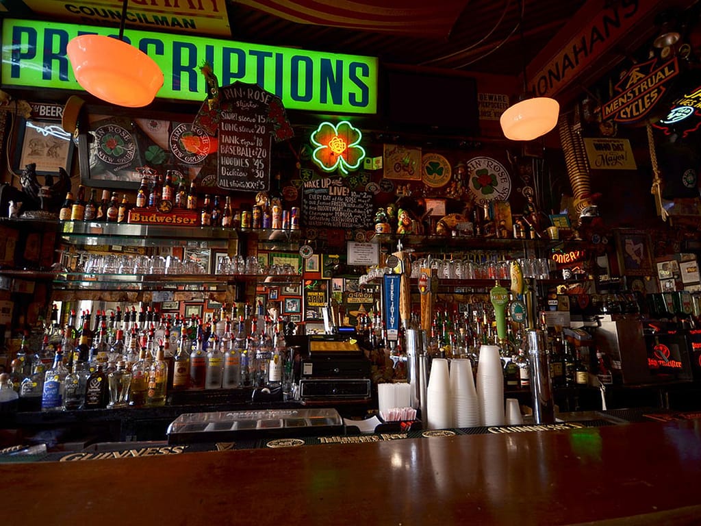 Busy bar interior with rows of liquor bottles, beer taps, glassware, and chalkboard menus behind the counter under neon signs.