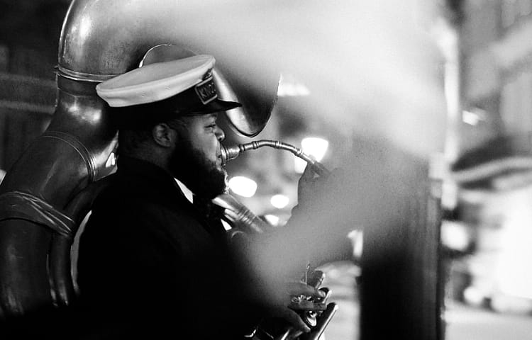 Black-and-white shot of a bearded man in a peaked cap playing a saxophone on a city street at night.