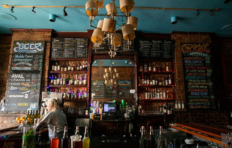 Bar interior with chalkboard menus, brick walls, and bottles on shelves beneath a large chandelier.